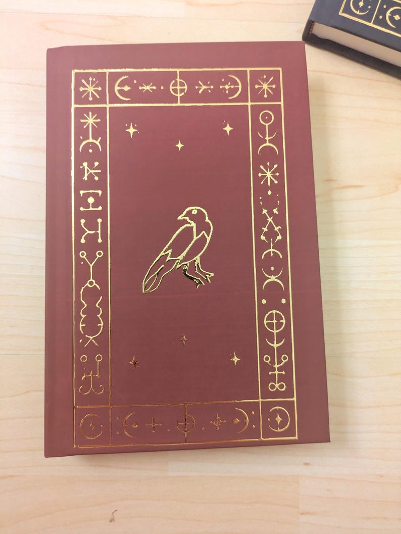 May include: A maroon book with gold foil accents. The cover features a gold raven illustration within a gold border of symbols and stars. The book is on a light wood surface.
