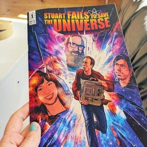 May include: A comic book titled Stuart Fails to Save the Universe, issue number 1, featuring a colorful cosmic background with shattered glass effects. The cover displays three illustrated characters and a central figure holding a technical device.