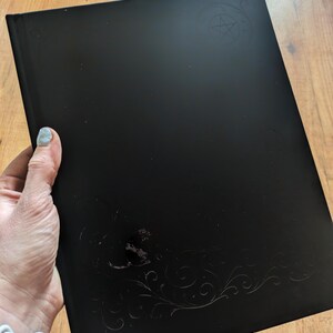 May include: A black journal with embossed decorative elements and a pentagram design. The journal is held in a person's hand. The cover has a glossy finish.