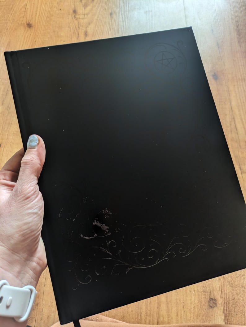 May include: A black journal with embossed decorative elements and a pentagram design. The journal is held in a person's hand. The cover has a glossy finish.
