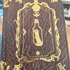 May include: A hardback book with a brown, textured cover and gold accents. The cover features a gold framed illustration of a bottle with a scroll inside. Gold anchor and decorative corner pieces add to the nautical theme.