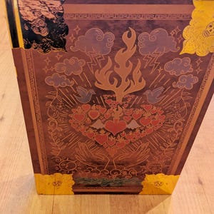 May include: A brown decorative box with gold accents and a detailed illustration. The illustration features a central flame, hearts, clouds, and hands. The box has gold corner protectors and a white top edge.