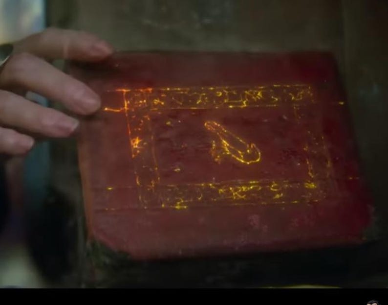 May include: A rectangular, reddish-brown book with glowing yellow symbols and a central design. The cover is being touched by a hand. The symbols and design appear to be illuminated.