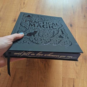 May include: A black book titled "PRACTICAL MAGIC" with a cat silhouette and decorative swirls. The book's spine reads "and fall in love whenever you can". A black ribbon bookmark is attached.