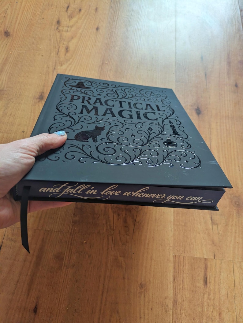May include: A black book titled "PRACTICAL MAGIC" with a cat silhouette and decorative swirls. The book's spine reads "and fall in love whenever you can". A black ribbon bookmark is attached.