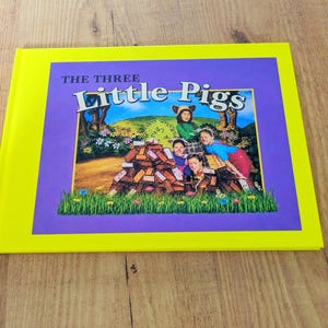 Puede incluir: Un libro infantil titulado The Three Little Pigs con un borde amarillo brillante y una cubierta morada. La ilustración central muestra a tres niños disfrazados de cerditos detrás de una pila de ladrillos marrones en un campo de hierba.