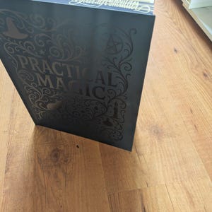 May include: A dark gray book titled "PRACTICAL MAGIC" with ornate gold lettering and illustrations. The book's cover features a witch's hat, stars, and a pentagram. The book is open, revealing the gold-edged pages.