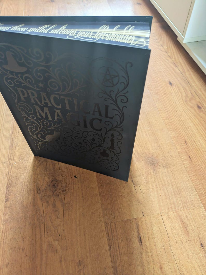 May include: A dark gray book titled "PRACTICAL MAGIC" with ornate gold lettering and illustrations. The book's cover features a witch's hat, stars, and a pentagram. The book is open, revealing the gold-edged pages.