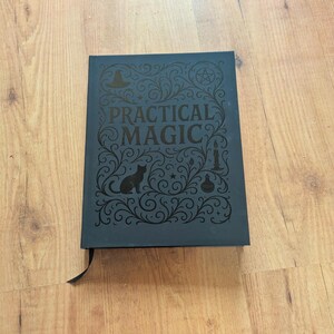May include: A dark blue book with the words "PRACTICAL MAGIC" in ornate lettering. The cover features a black cat silhouette, a witch's hat, a pentagram, a candle, and a potion bottle. A black ribbon bookmark is visible.