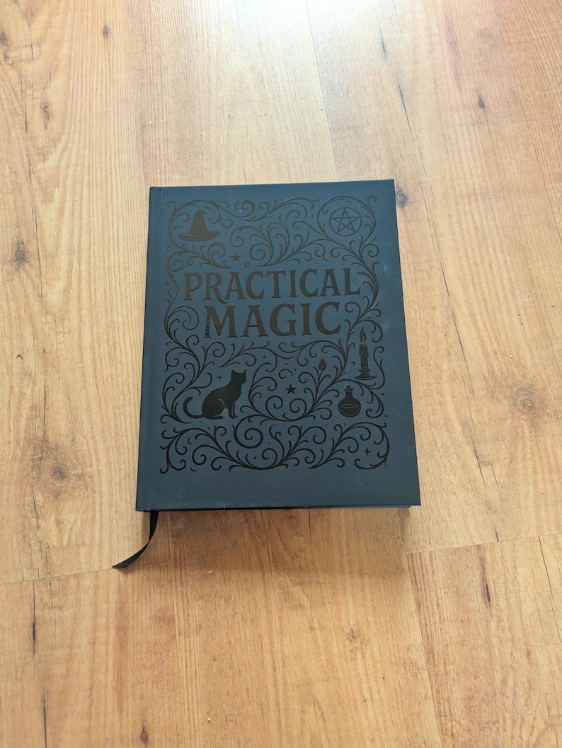 May include: A dark blue book with the words "PRACTICAL MAGIC" in ornate lettering. The cover features a black cat silhouette, a witch's hat, a pentagram, a candle, and a potion bottle. A black ribbon bookmark is visible.