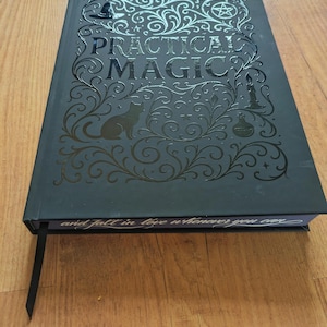 May include: A black hardcover book titled "Practical Magic" with decorative silver embossed designs, including a cat silhouette, a witch's hat, and a pentagram. The book has a black ribbon bookmark and the text "and fall in love whenever you can".