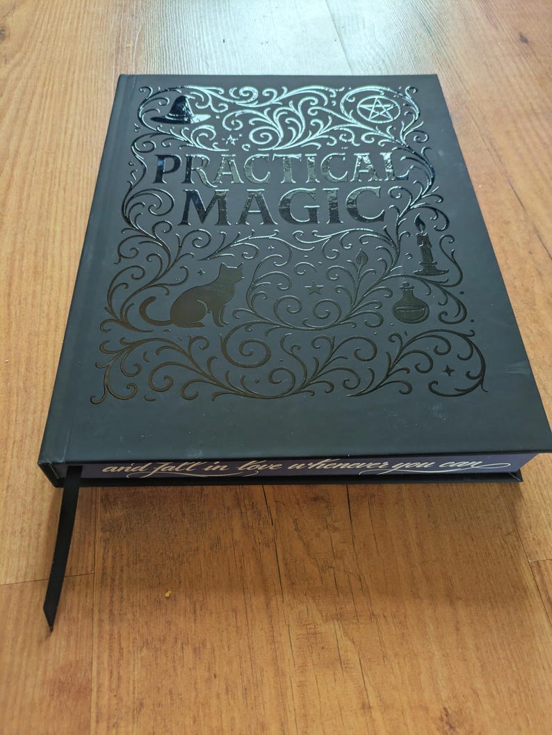 May include: A black hardcover book titled "Practical Magic" with decorative silver embossed designs, including a cat silhouette, a witch's hat, and a pentagram. The book has a black ribbon bookmark and the text "and fall in love whenever you can".