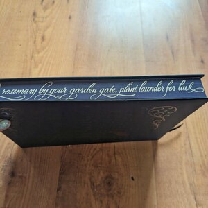 May include: A dark blue book with gold lettering that reads "rosemary by your garden gate, plant launder for luck." The book has gold decorative accents and a black ribbon bookmark. The book is on a wooden surface.