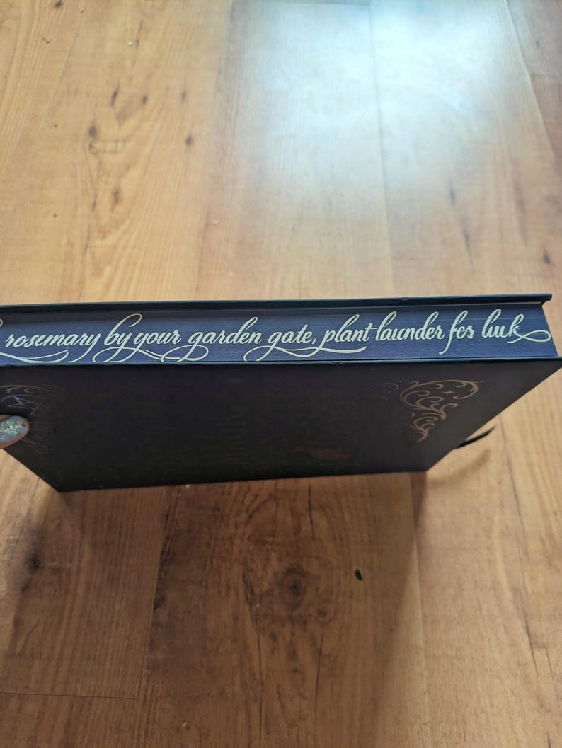 May include: A dark blue book with gold lettering that reads "rosemary by your garden gate, plant launder for luck." The book has gold decorative accents and a black ribbon bookmark. The book is on a wooden surface.