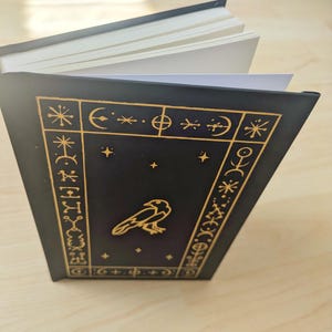 May include: A black journal with a gold raven illustration and celestial symbols. The cover features a gold border with stars, moons, and other symbols. The pages are white and the journal is open.