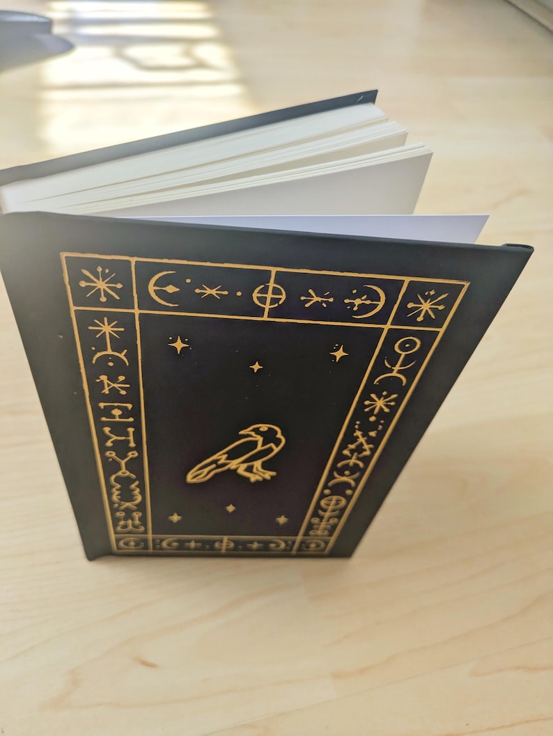 May include: A black journal with a gold raven illustration and celestial symbols. The cover features a gold border with stars, moons, and other symbols. The pages are white and the journal is open.