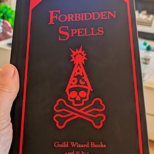 May include: A black book with the title "Forbidden Spells" in red lettering. The cover design includes a red skull and crossbones, a wizard's hat, and a starburst. The text "Guild Wizard Books 43rd Edition" is also present.