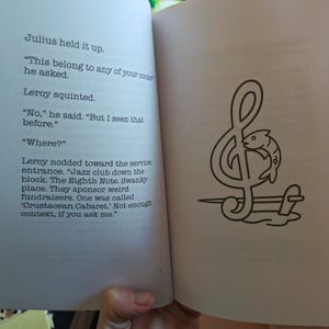 Pu&ograve; includere: Un libro aperto con testo e un'illustrazione in bianco e nero. L'illustrazione &egrave; una nota musicale con un pesce all'interno. Il testo recita: "Julius lo sollev&ograve;. 'Questo appartiene a qualcuno dei tuoi cuochi?' chiese."