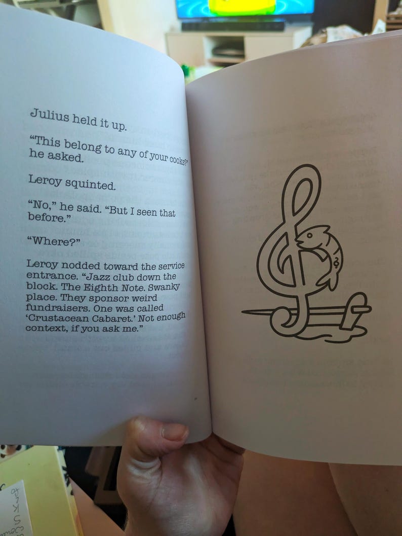 Pu&ograve; includere: Un libro aperto con testo e un'illustrazione in bianco e nero. L'illustrazione &egrave; una nota musicale con un pesce all'interno. Il testo recita: "Julius lo sollev&ograve;. 'Questo appartiene a qualcuno dei tuoi cuochi?' chiese."