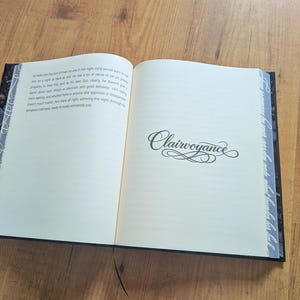 May include: An open book with the word "Clairvoyance" in elegant script on the right page. The left page contains printed text. The book has a black cover with decorative edges and a black bookmark.