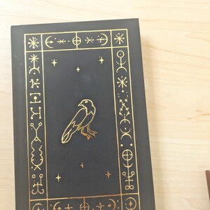 May include: A black book with gold foil accents. The cover features a gold raven illustration and a decorative border with celestial symbols. The book is closed and resting on a light wood surface.
