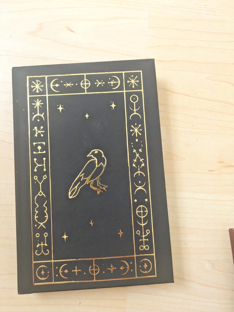 May include: A black book with gold foil accents. The cover features a gold raven illustration and a decorative border with celestial symbols. The book is closed and resting on a light wood surface.