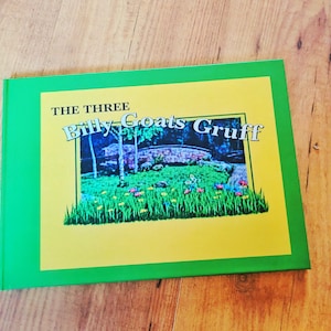 May include: A rectangular book with a bright green border and a yellow centre panel. The cover features the text THE THREE Billy Goats Gruff above an illustration of a stone bridge over a grassy field with small colourful flowers and trees.