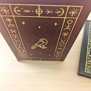 May include: A maroon book with gold embossed designs and a raven illustration. The cover features geometric shapes, stars, and symbols. The book's spine is visible, and another book is partially seen in the background.