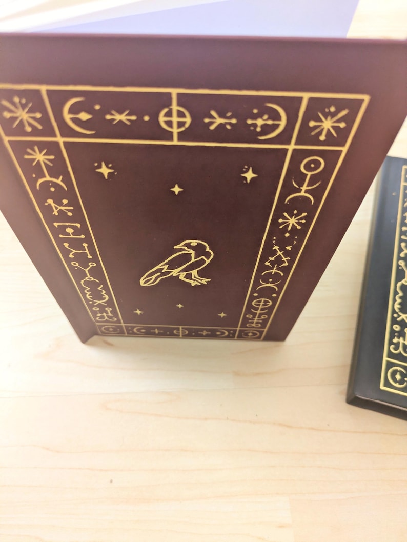 May include: A maroon book with gold embossed designs and a raven illustration. The cover features geometric shapes, stars, and symbols. The book's spine is visible, and another book is partially seen in the background.