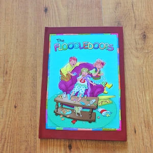 Puede incluir: Un libro de tapa dura titulado The FLOODLEDOOPS con una cubierta color granate. La ilustración frontal muestra a tres niños sentados en un sofá morado comiendo, con una mesa de centro frente a ellos llena de comida, migas y un gato pequeño.