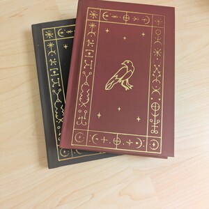 May include: Two hardcover books with gold foil designs. One book is burgundy with a gold raven illustration and celestial symbols. The other book is black with gold celestial symbols. The books are on a light wood surface.