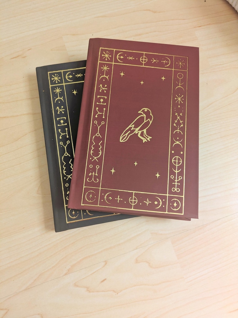 May include: Two hardcover books with gold foil designs. One book is burgundy with a gold raven illustration and celestial symbols. The other book is black with gold celestial symbols. The books are on a light wood surface.