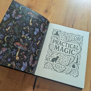 May include: An open book with a black cover, displaying two pages. The left page features a dark background with butterflies, mushrooms, and flowers. The right page reads "PRACTICAL MAGIC" with decorative swirls, a cat silhouette, and a witch's hat.