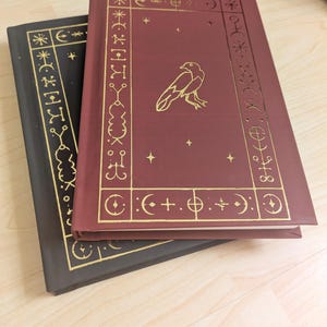 May include: Two hardcover books, one black and one burgundy, stacked on top of each other. The burgundy book features a gold raven illustration and gold border designs. The black book has gold border designs. Both books have a rectangular shape.