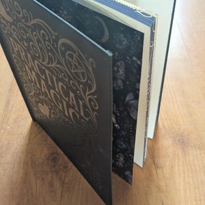 May include: A dark book titled "Practical Magick" with gold lettering and decorative accents. The book is open, revealing pages with illustrations and text. The edges of the pages are decorated with gold trim. The book is on a wooden surface.