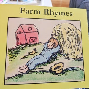 Puede incluir: Una portada de libro titulada Farm Rhymes con un borde amarillo. La ilustración muestra a una persona con ropa azul descansando contra un gran pajar junto a un granero rojo, un pájaro pequeño y un sombrero sobre la hierba verde.