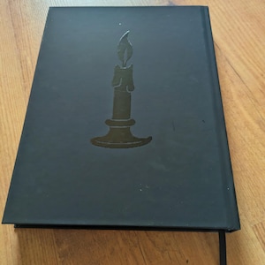 May include: Black journal with a debossed candle design on the cover. The candle is centered and has a flame. A black ribbon bookmark is visible. The journal is closed and resting on a wooden surface.