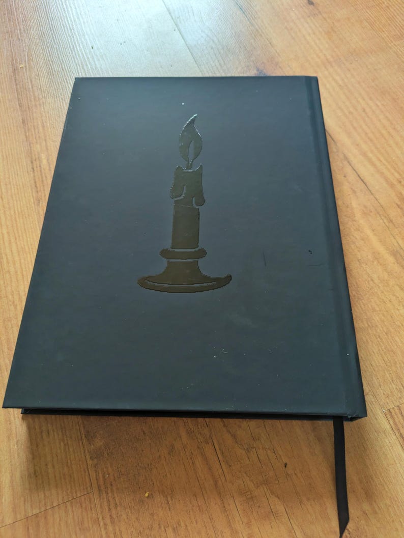 May include: Black journal with a debossed candle design on the cover. The candle is centered and has a flame. A black ribbon bookmark is visible. The journal is closed and resting on a wooden surface.