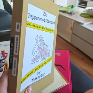 Pu&ograve; includere: Un libro con copertina color beige intitolato "The Pepperwood Chronicles" con un'etichetta bianca raffigurante un'aragosta e la scritta "POLICE LINE - DO NOT CROSS". &Egrave; visibile anche il nome dell'autore, Nick Miller. Altri libri sono sullo sfondo.