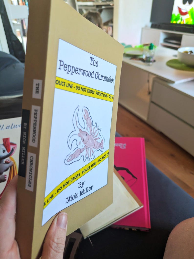Pu&ograve; includere: Un libro con copertina color beige intitolato "The Pepperwood Chronicles" con un'etichetta bianca raffigurante un'aragosta e la scritta "POLICE LINE - DO NOT CROSS". &Egrave; visibile anche il nome dell'autore, Nick Miller. Altri libri sono sullo sfondo.