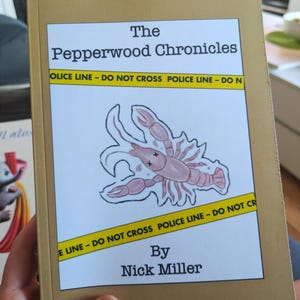 Può includere: Un libro intitolato "The Pepperwood Chronicles" con una copertina dorata. La copertina presenta un'illustrazione di aragosta rosa e nastro della polizia giallo. Il nome dell'autore, Nick Miller, è stampato in basso.