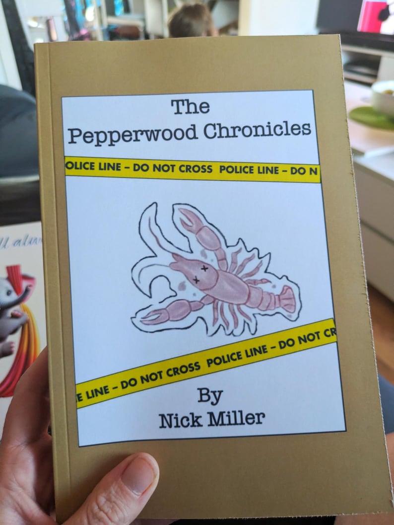 Pu&ograve; includere: Un libro intitolato "The Pepperwood Chronicles" con una copertina dorata. La copertina presenta un'illustrazione di aragosta rosa e nastro della polizia giallo. Il nome dell'autore, Nick Miller, &egrave; stampato in basso.