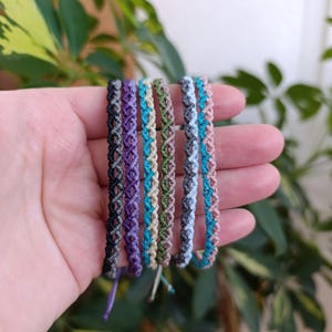 May include: A hand displays six macrame bracelets in various colors, including black, purple, green, blue, and pink. The bracelets have a woven design and are made of cord. The background features green foliage.