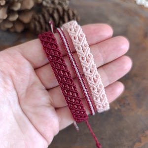 Può includere: Sono presentati tre braccialetti in macramè nei toni del rosso e del rosa. I braccialetti presentano intricati motivi intrecciati. Uno è bordeaux scuro, un altro è rosa chiaro e il terzo combina rosso e bianco. I braccialetti sono fatti a mano.