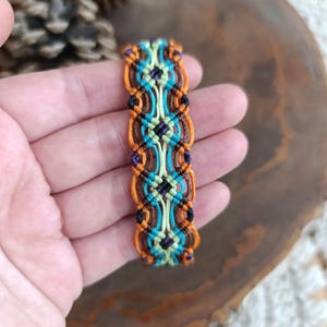 May include: A handmade macrame bracelet featuring a repeating pattern of orange, teal, brown, and light green threads. The bracelet has a rectangular shape with black and purple accents. The bracelet is approximately 1 inch wide.