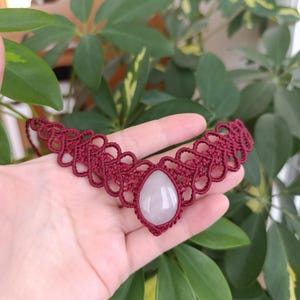 Puede incluir: Un collar de macramé burdeos hecho a mano con un colgante de piedra preciosa rosa pálido en forma de lágrima. El intrincado trabajo de nudos forma un delicado diseño de collar. El collar se muestra sobre un fondo de follaje verde.