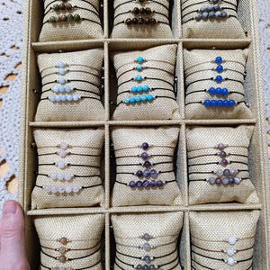 Puede incluir: Una exhibición de pulseras de cuentas en una caja dividida. Cada pulsera presenta una serie de pequeñas cuentas de colores ensartadas en un cordón negro. Las cuentas varían en color, incluyendo verde, marrón, azul y morado. Las pulseras están dispuestas sobre pequeñas almohadas beige.