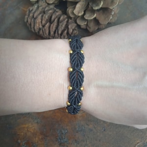 Könnte beinhalten: Schwarzes geflochtenes Armband mit goldenen Perlen. Das Armband ist mit einem gewebten Muster gefertigt und hat ein schlichtes Design.