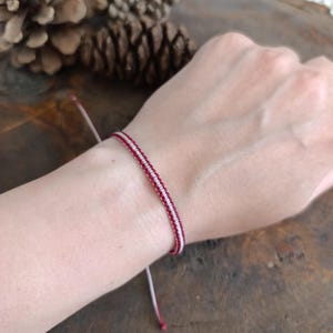 Puede incluir: Pulsera hecha a mano con un diseño tejido con bandas alternas rojas y blancas. La pulsera se asegura con cordones ajustables. La pulsera se muestra en una muñeca, mostrando los intrincados detalles y el contraste de color.