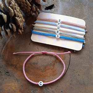 Könnte beinhalten: Ein Set aus fünf Freundschaftsarmbändern mit verschiedenen farbigen Schnüren und weißen Buchstabenperlen, die "SPDBS" buchstabieren. Ein rosafarbenes Armband mit einer weißen Buchstabenperle, die "C" buchstabiert, ist ebenfalls abgebildet.